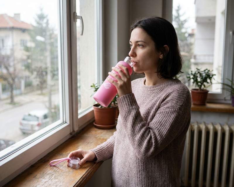 Жена до прозореца пие от розовата стъклена бутилка Hygea Water